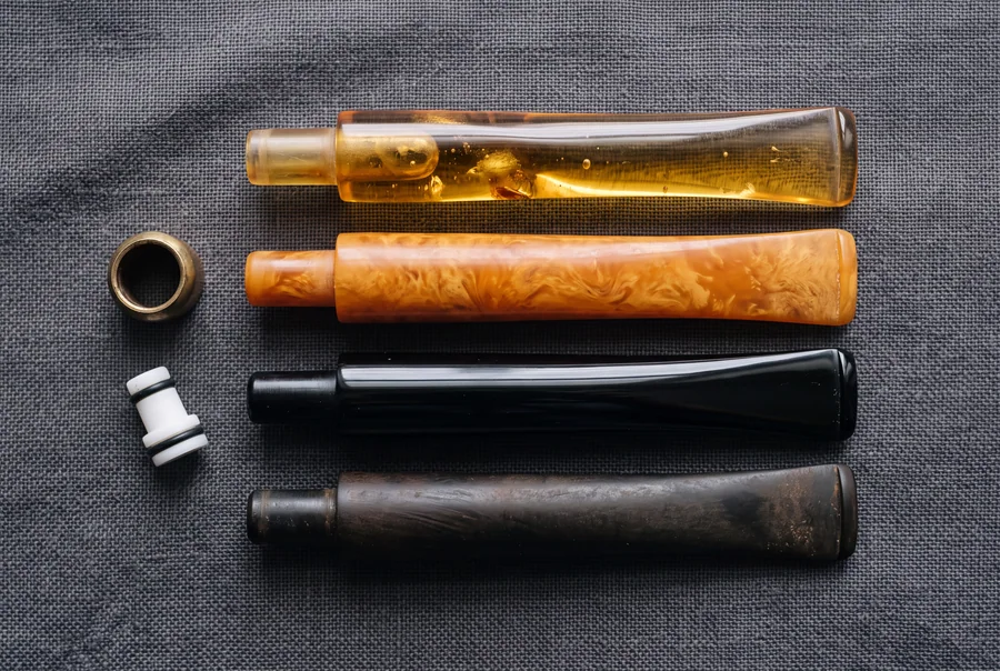 Four pipe stems laid in a row on grey linen: translucent Baltic amber, cloudy pressed amber, glossy black acrylic and dulled vulcanite